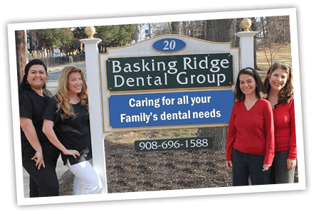 The image shows three individuals posing in front of a sign for Basking Ridge Dental Group, which includes a woman standing on the left side of the sign, two women standing on the right side of the sign, and a sign with text that reads  BASKING RIDGE DENTAL GROUP  at the top, followed by  CARE FOR ALL YOUR DENTAL NEEDS  in smaller font, and  1234 Main Street  below. The style of the image is a casual photograph with an overlaid text watermark that reads  www.yourwebsite.com.