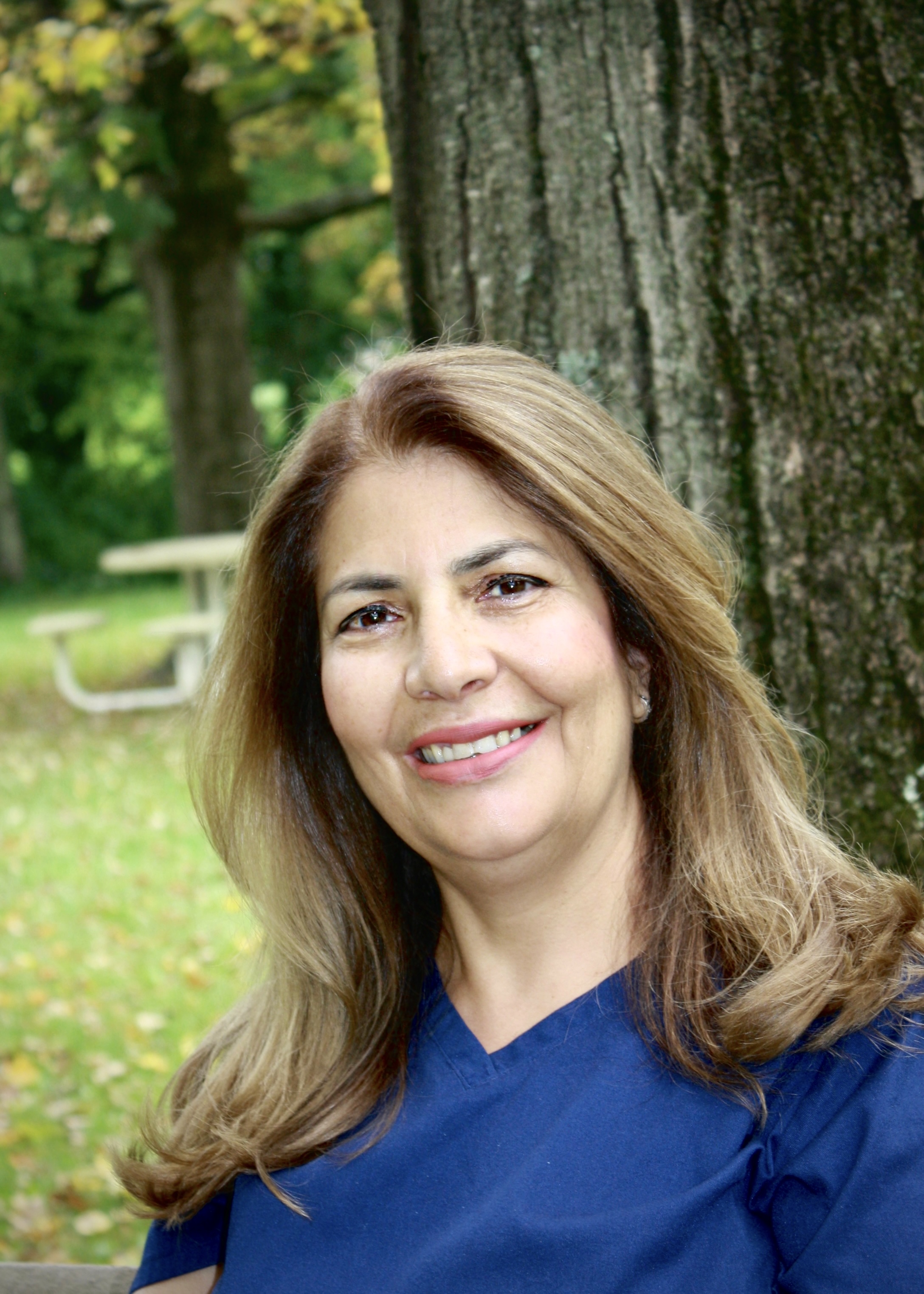 The image features a woman wearing a white coat over a blue shirt, posing for a portrait with a smile against a backdrop of a park setting with trees and a bench.