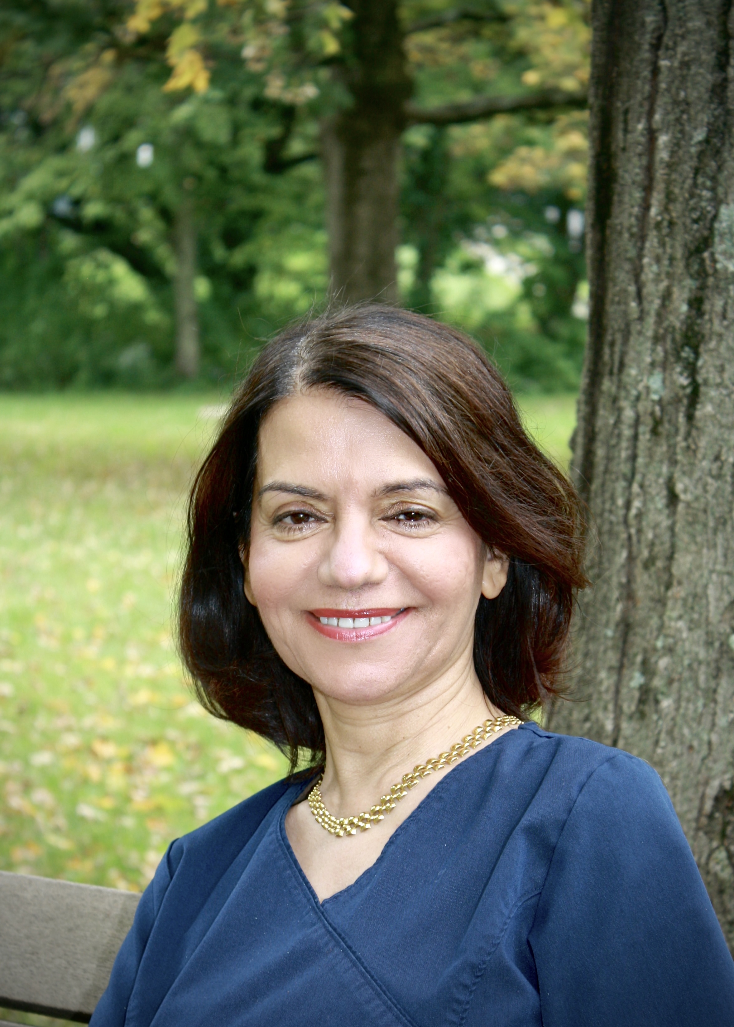 Woman with dark hair and a smile, wearing a blue shirt, seated outdoors on a bench.