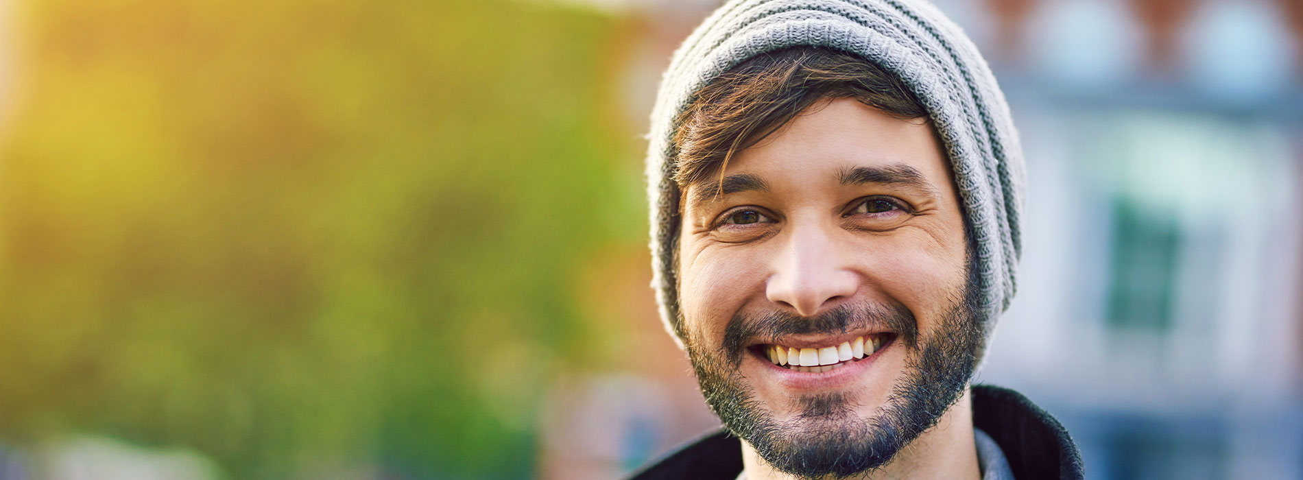 The image features a young man with a beard and mustache, smiling broadly at the camera, wearing a beanie and sunglasses, set against a blurred cityscape background.