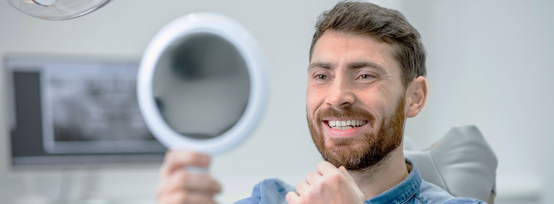 A man holding a round mirror up to his face in front of a backdrop with medical equipment.