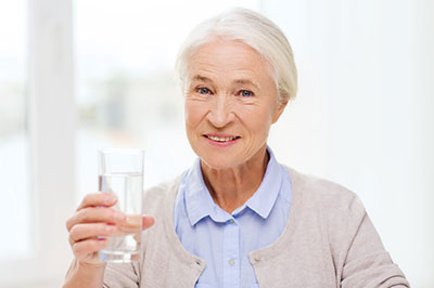 The image shows an older woman holding a glass of water, smiling at the camera, with her right hand raised slightly above the glass.