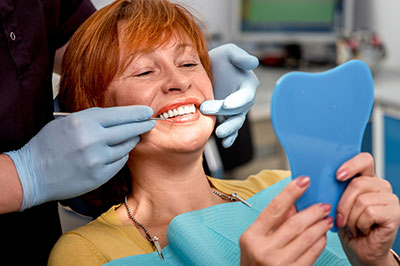 The image depicts a woman sitting in a dental chair with a smile, holding a blue dental impression tray while wearing a white surgical mask, with a dental professional adjusting her teeth in the background.