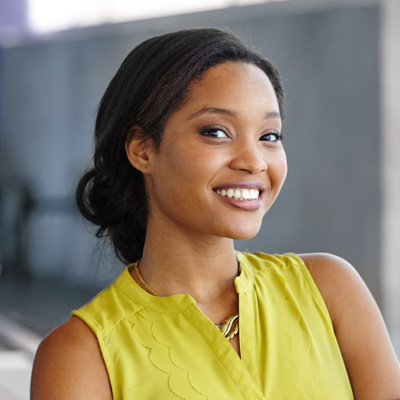 The image depicts a woman with dark hair smiling at the camera, wearing a yellow top and standing against a light-colored background.