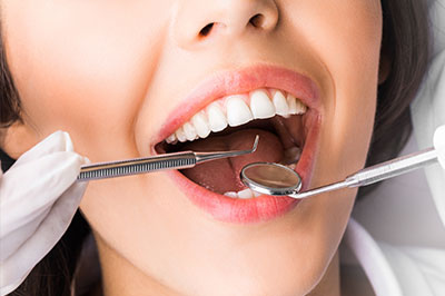 A woman wearing a white lab coat is sitting at a dental chair with her mouth open, receiving dental care from a professional holding dental instruments.